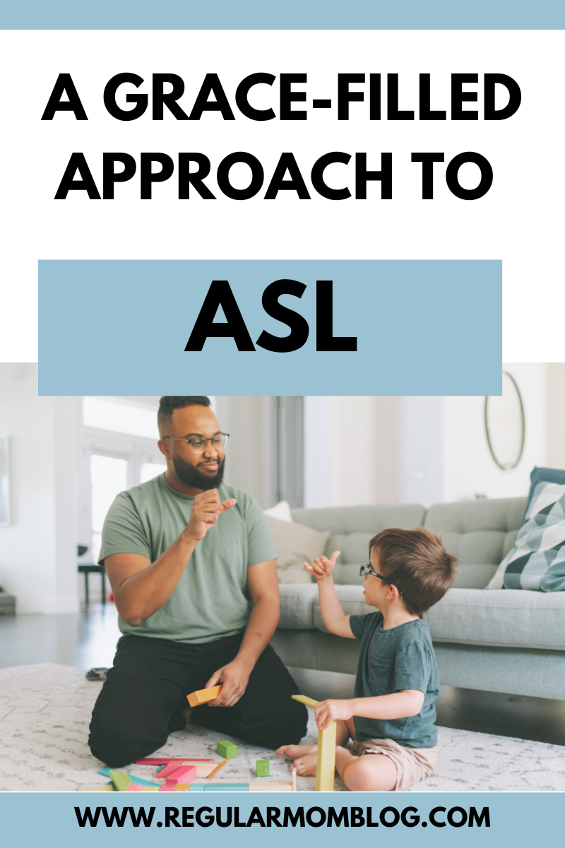 A black man sits on the living room floor with his white sone with glasses. They are playing with toys and the man is teaching his son ASL.