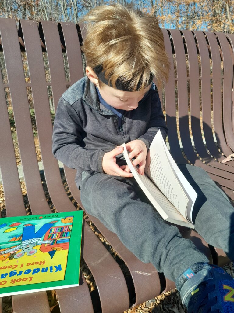 a boy practices speech goals at home. he sits on a park bench and is reading a book with another book sitting next to him.
