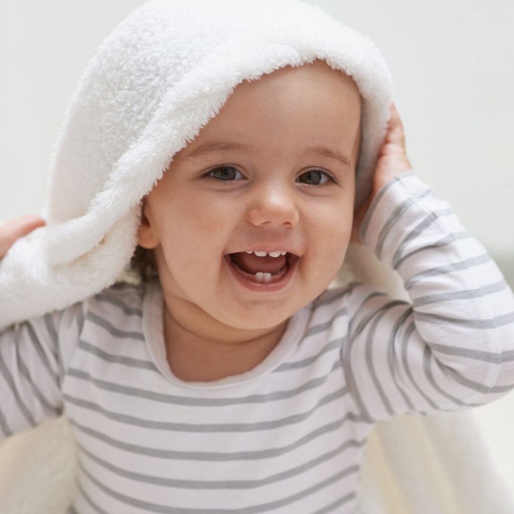 A baby girl sits up on a bed and has a white blanket over her head. She is playing peek a boo sensory activity for babies.