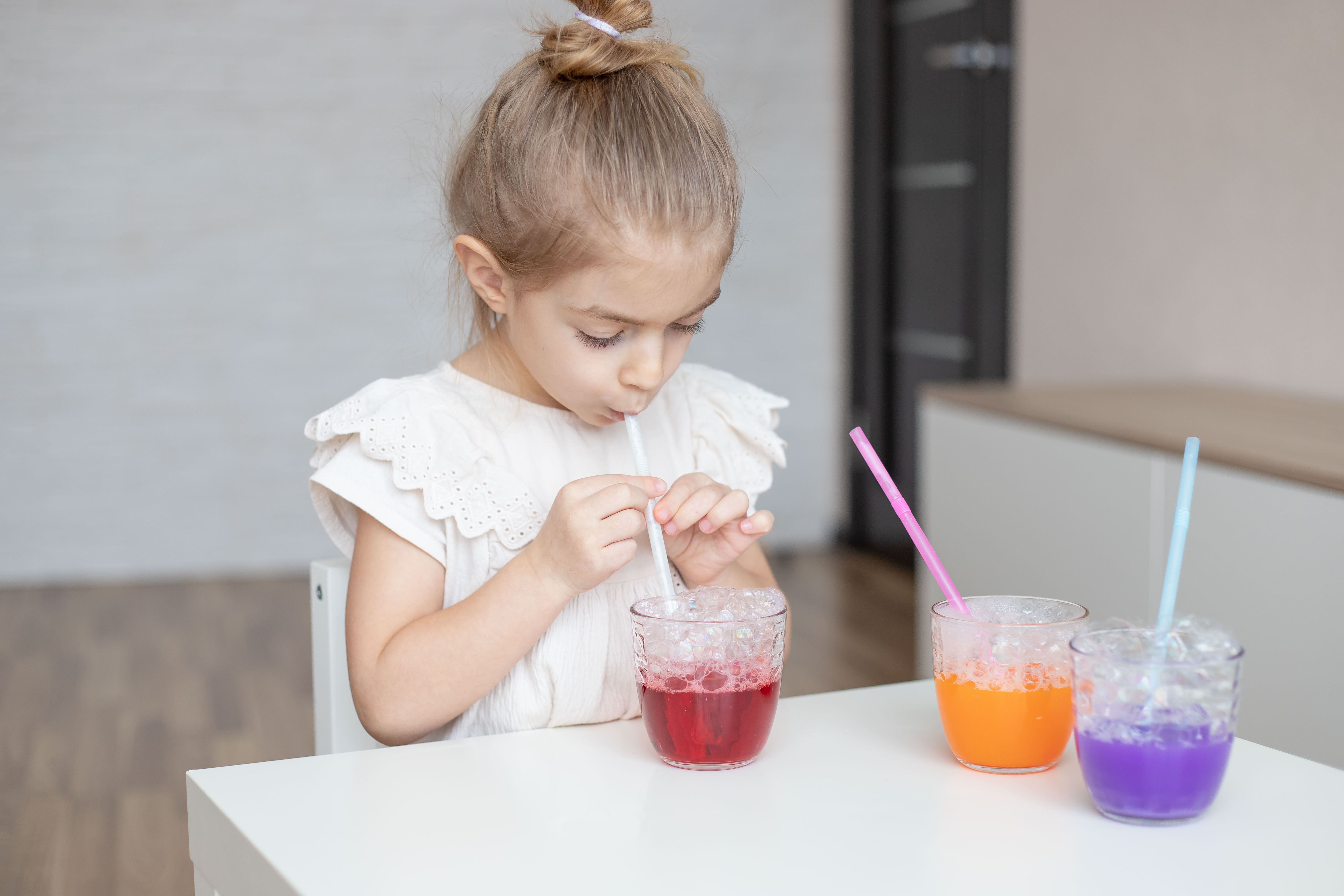 a girl sits at a desk with different colored cups in front of her and she blows into the cups with straws for a bubble activity