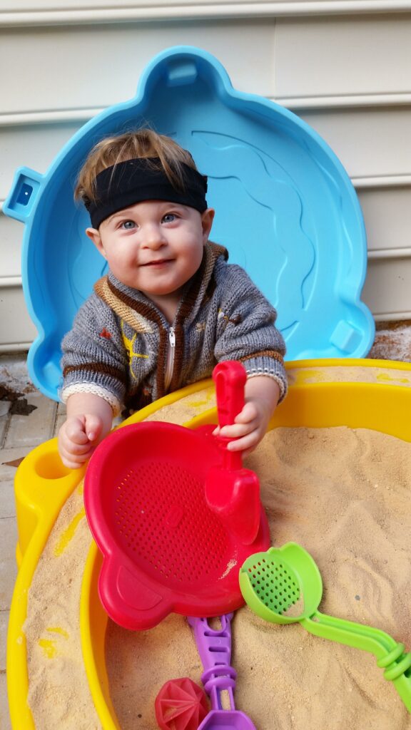 A toddler boy stands behind a sand box and plays sand activities while holding a shovel
