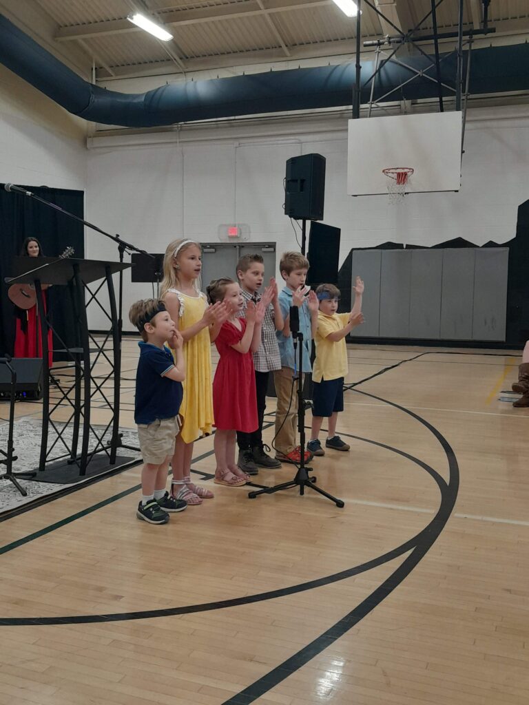 a group of kids standing in a gym auditorium singin a song and doing ASL signs to the song.
