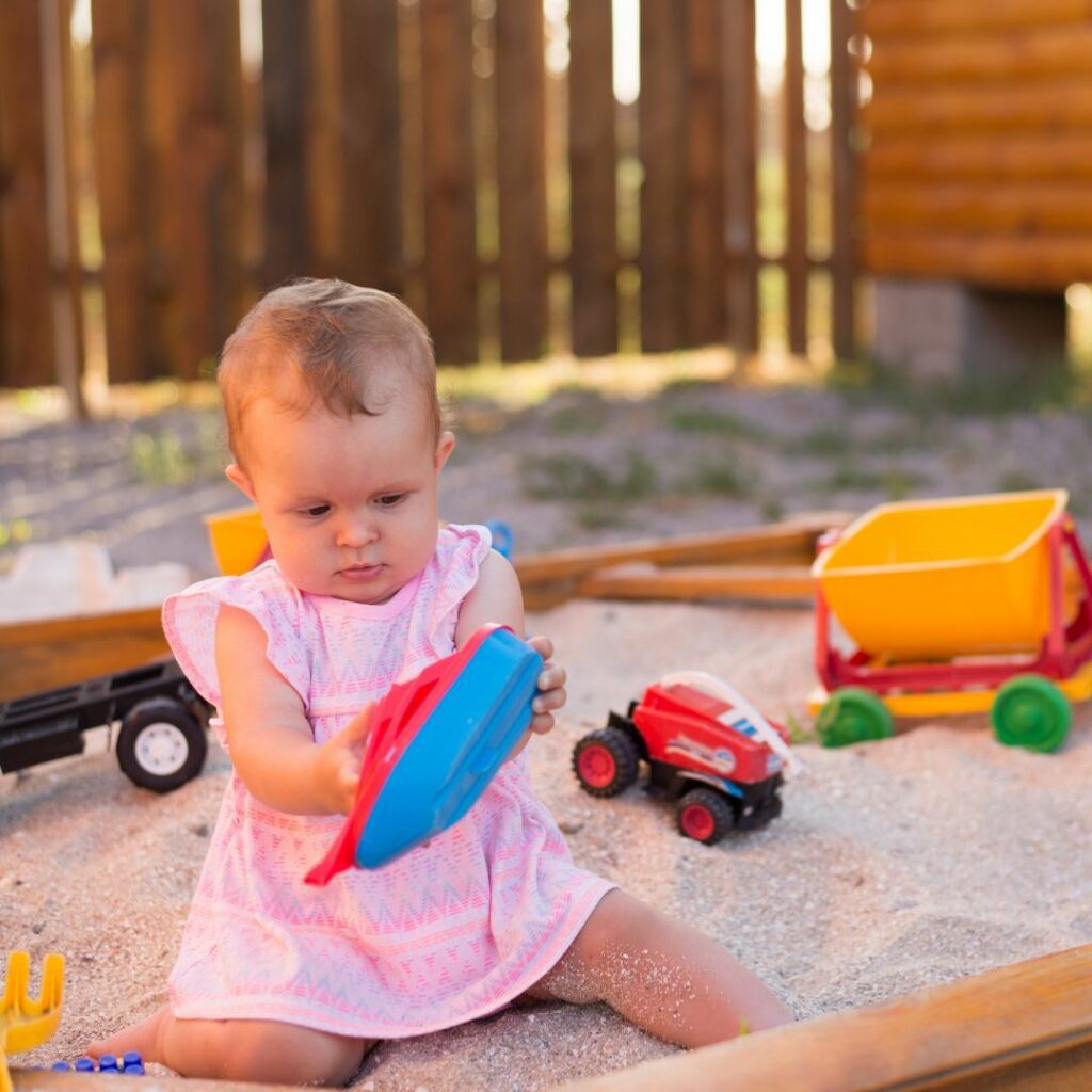 a baby sits in a sandbox at dusk and plays with sand toys