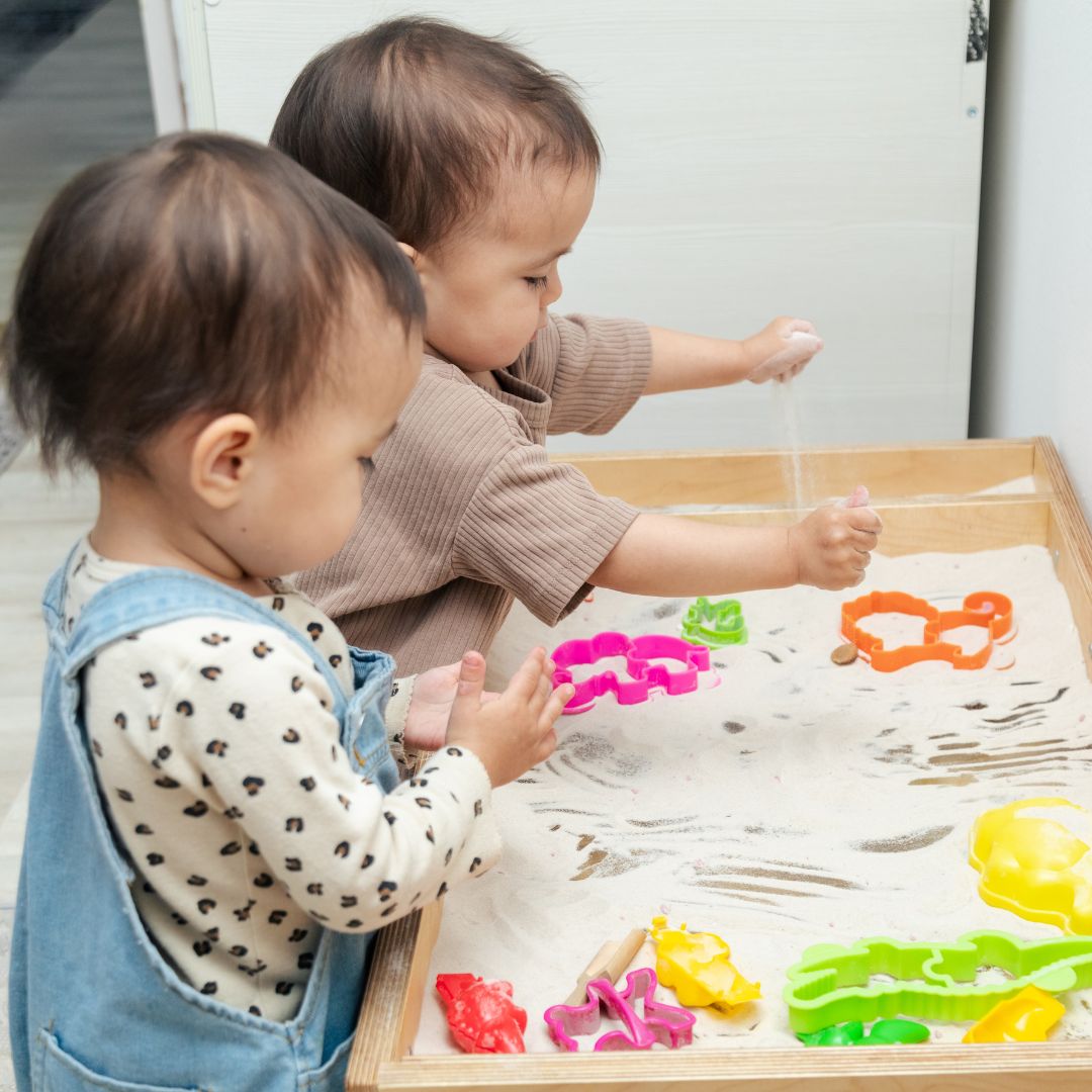 two babies stand at a sand tabl e and play sensory sand play