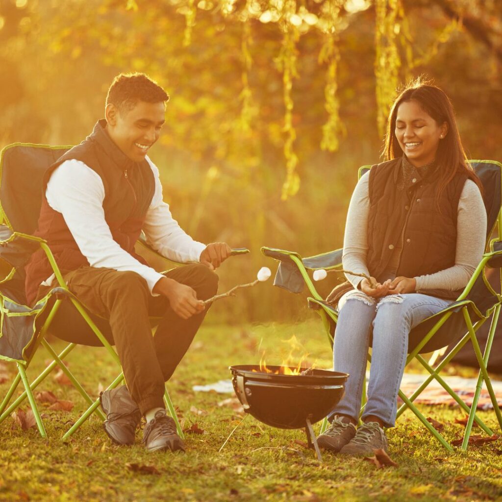 a couple sit in camping chairs in front of a fire and roast marshmallows