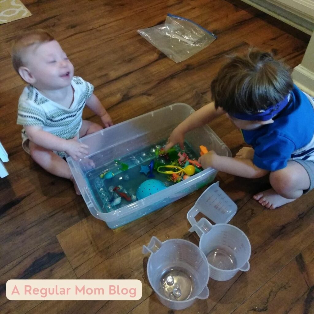 a baby and a toddler play with a bin of water in a sensory game.