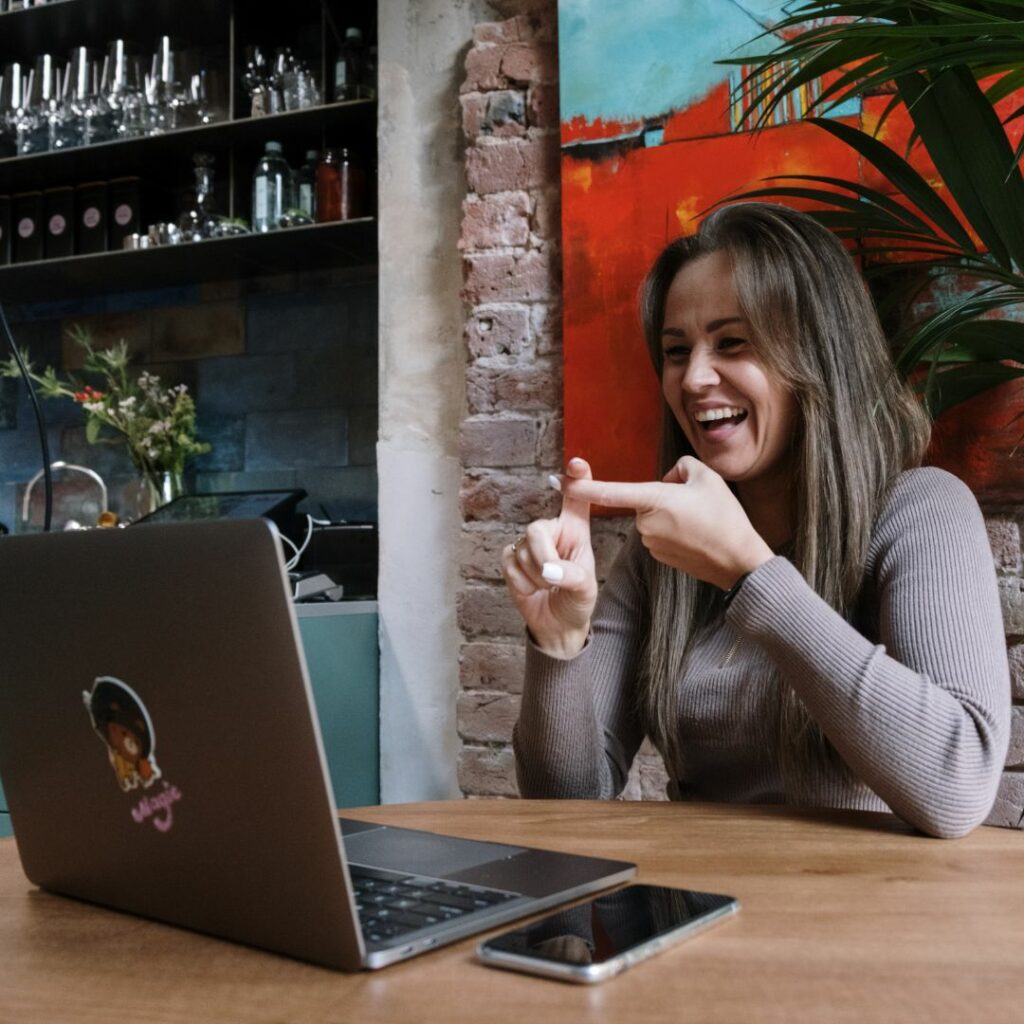 a woman sits at a table in a coffee shop with a laptop in front of her and learns ASL