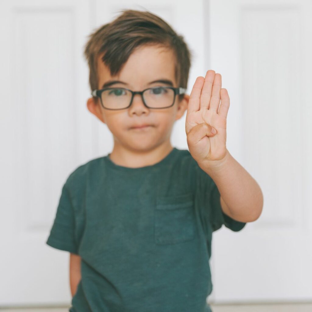 a boy with glasses sits on the floor and signs the letter B with his hands for an ASL sign