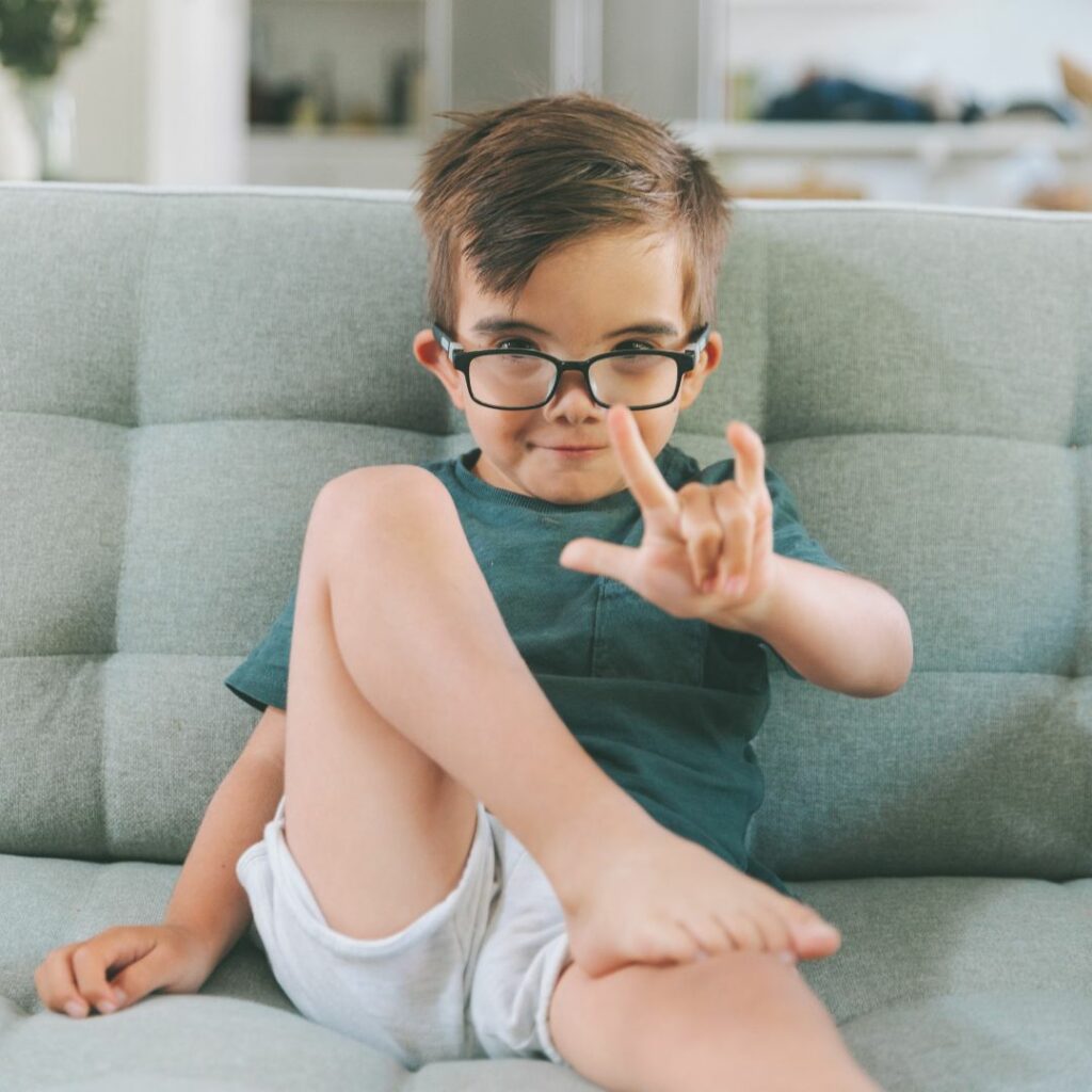 a toddler boy with glasses sits on a sofa and does the i love you asl sign