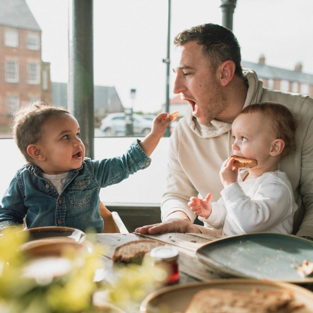 a father holds his baby and eats with his other baby at the table for a taste sensory activity