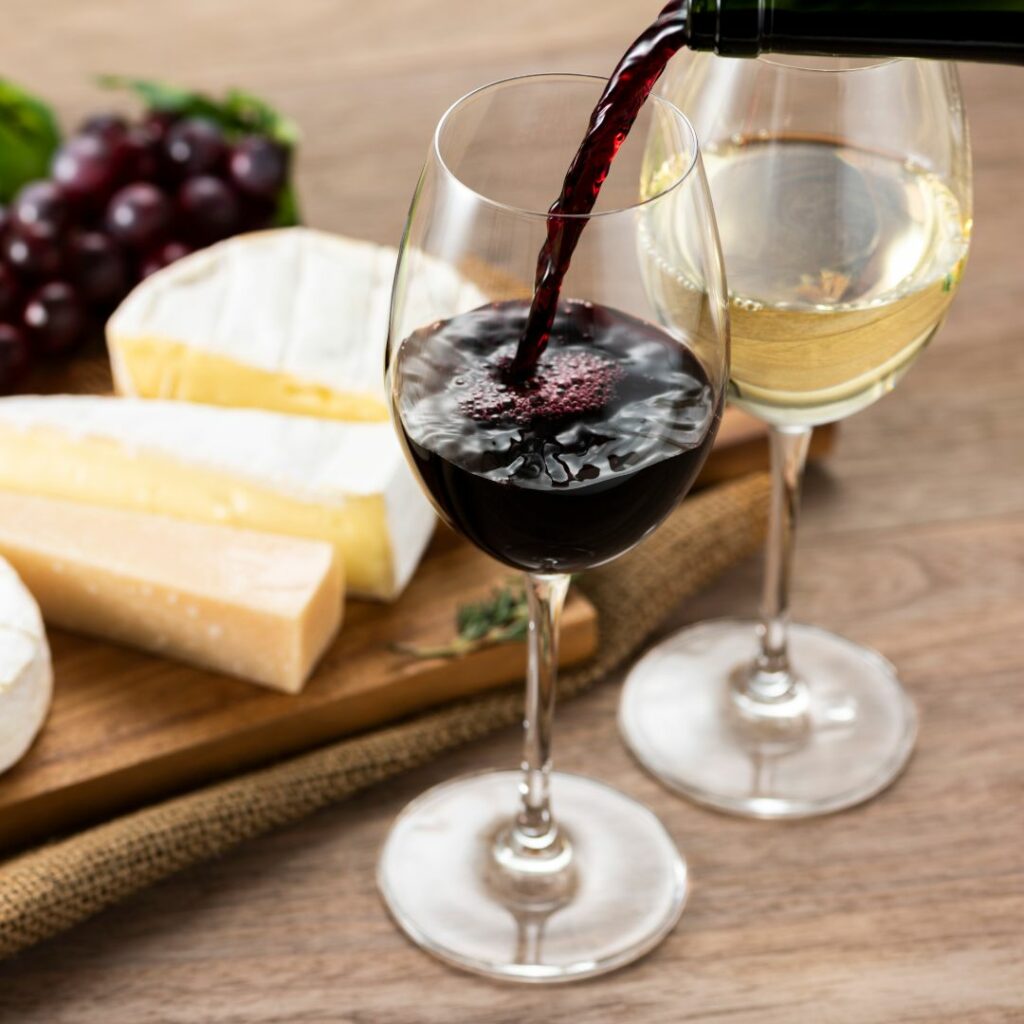 two glasses of wine sit on a table with cheese in the background, and someone it pouring the red wine
