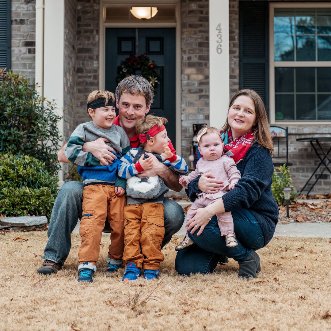 two parents sit with their children outside of their house they have three children, two boys and one girl on their knees in front of them and they are all smiling