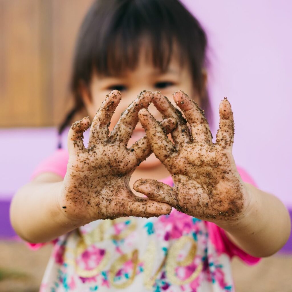 a toddler girl shows off her dirty and muddy hands after a nature sensory activity