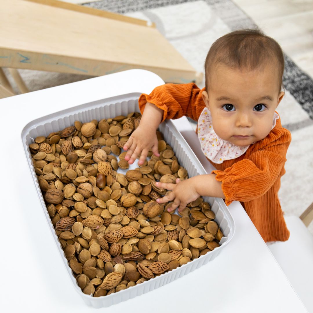 a 12 month old baby stands at a table and plays with a sensory bin filled with nuts