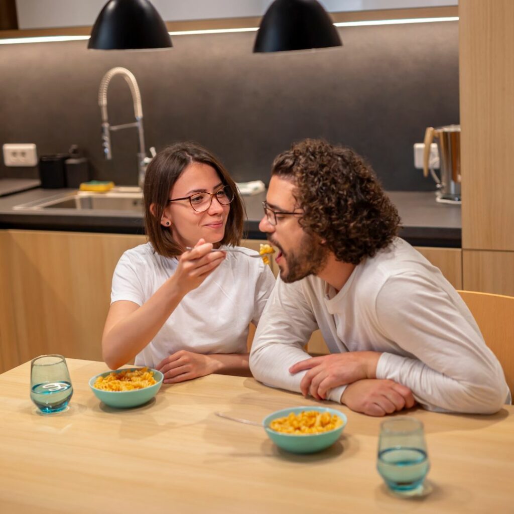 a couple sits at a table in their kitchen and eat macaroni and cheese together for a dinner date night at home