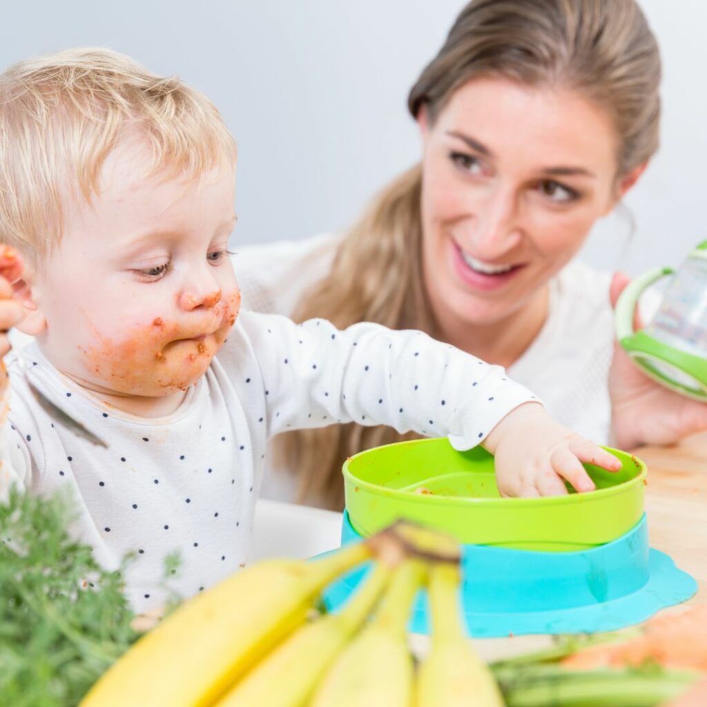 a mother sits beside her baby who is in a high chair and the baby tries all different types of foods for a high chair sensory play activity