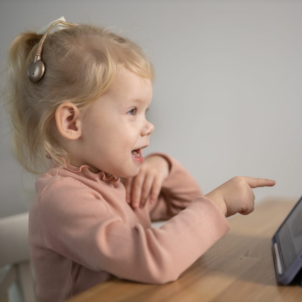 a little girl wearing gold cochlear implants sits at a table and watches a computer screen.