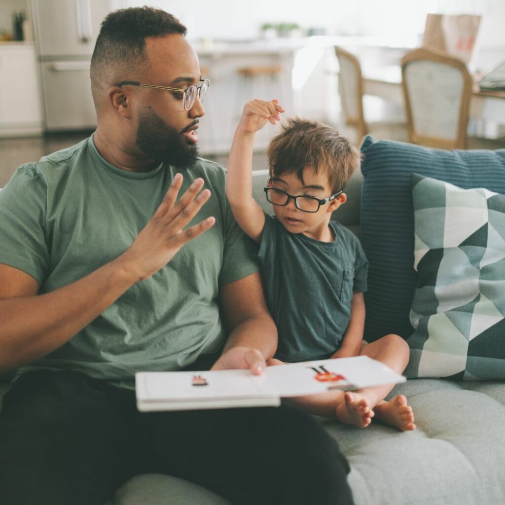 a dad and son sit on the sofa and the dad teaches sign language to his toddler son