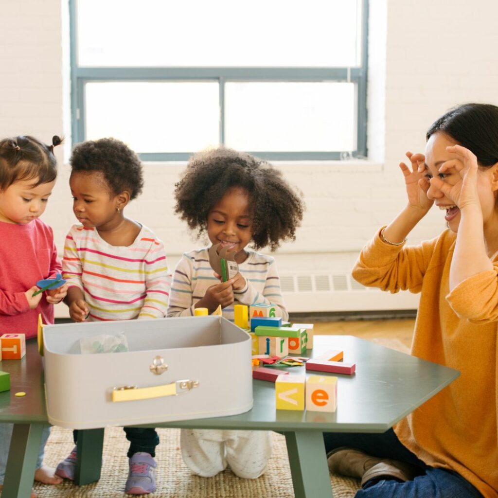 a teacher sits at a long table with her toddler students and tries to do a sign for I see