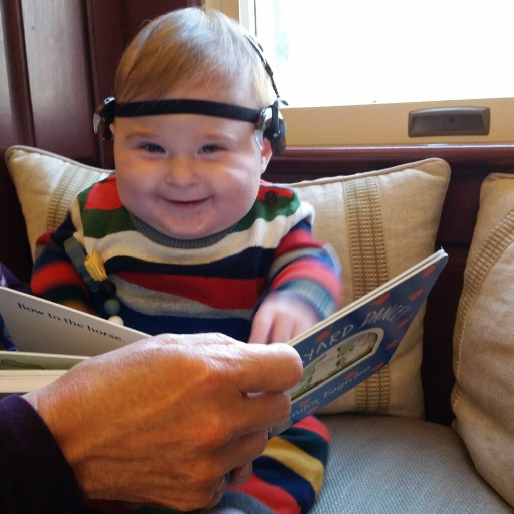 a one year old wears cochlear implants and sits on the sofa and is looking at a book