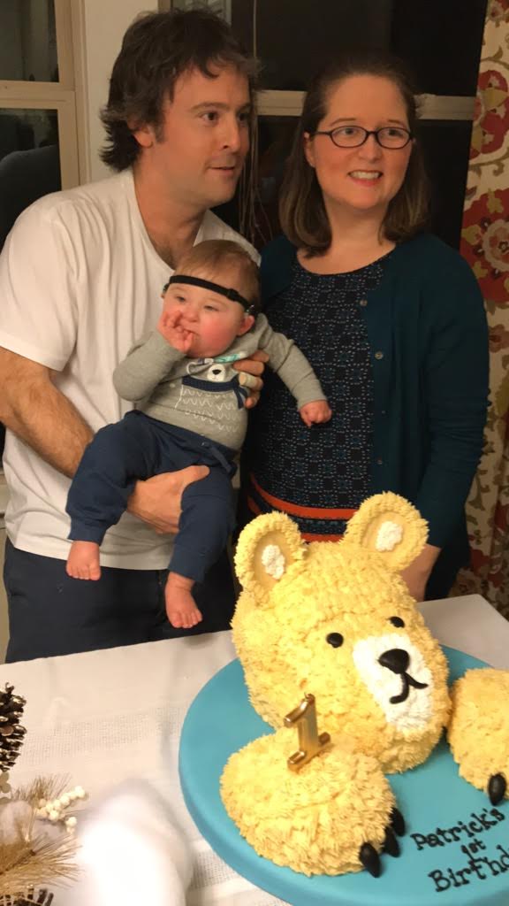 two parents hold their one year old baby and stand behind a table with a bear cake on it. the baby wears cochlear implants