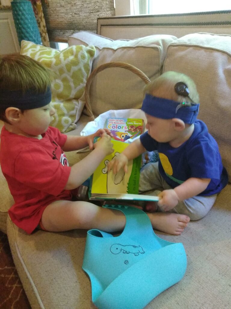 a 2year old boy and a 1 year old boy sit on the sofa together with an Easter basket in the middle of them. They are both wearing cochlear implants.
